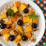 A top view of a breakfast bowl of steel-cut oats topped with fresh mango, grapes, walnuts, and mint leaves.