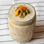 ar of coconut mango overnight oats topped with mango slices and pumpkin seeds on a striped cloth background.