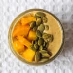 Overhead view of a mango peanut butter smoothie topped with fresh mango chunks and pumpkin seeds, served in a glass jar on a white textured background.
