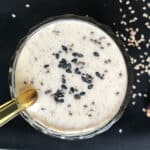 Creamy tahini banana smoothie in a glass, topped with black sesame seeds and chia seeds, with dates and sesame scattered on a dark background.