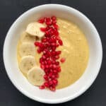 Creamy mango banana smoothie bowl garnished with fresh banana slices and pomegranate arils.