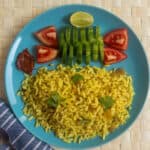 A blue plate with puffed rice upma garnished with coriander, served with sliced cucumber, tomato wedges, half a lime, and a side of pickle on a woven mat.